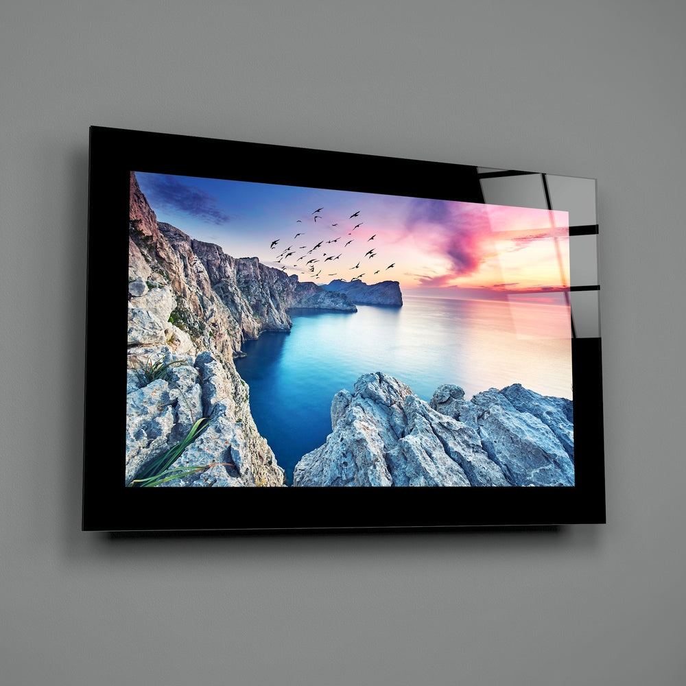 Black-framed glass wall art displaying a coastal sunset scene with rocky cliffs, blue water, and flying birds, mounted on gray wall.