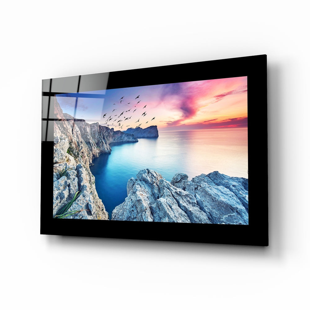 Black-framed glass wall art displaying a coastal sunset scene with rocky cliffs, turquoise water, pink sky, and flying birds.