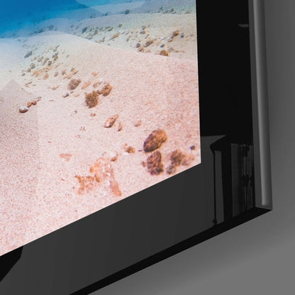Close-up angled view of glass wall art displaying an underwater seafloor with sandy bottom and scattered brown rocks against blue water.