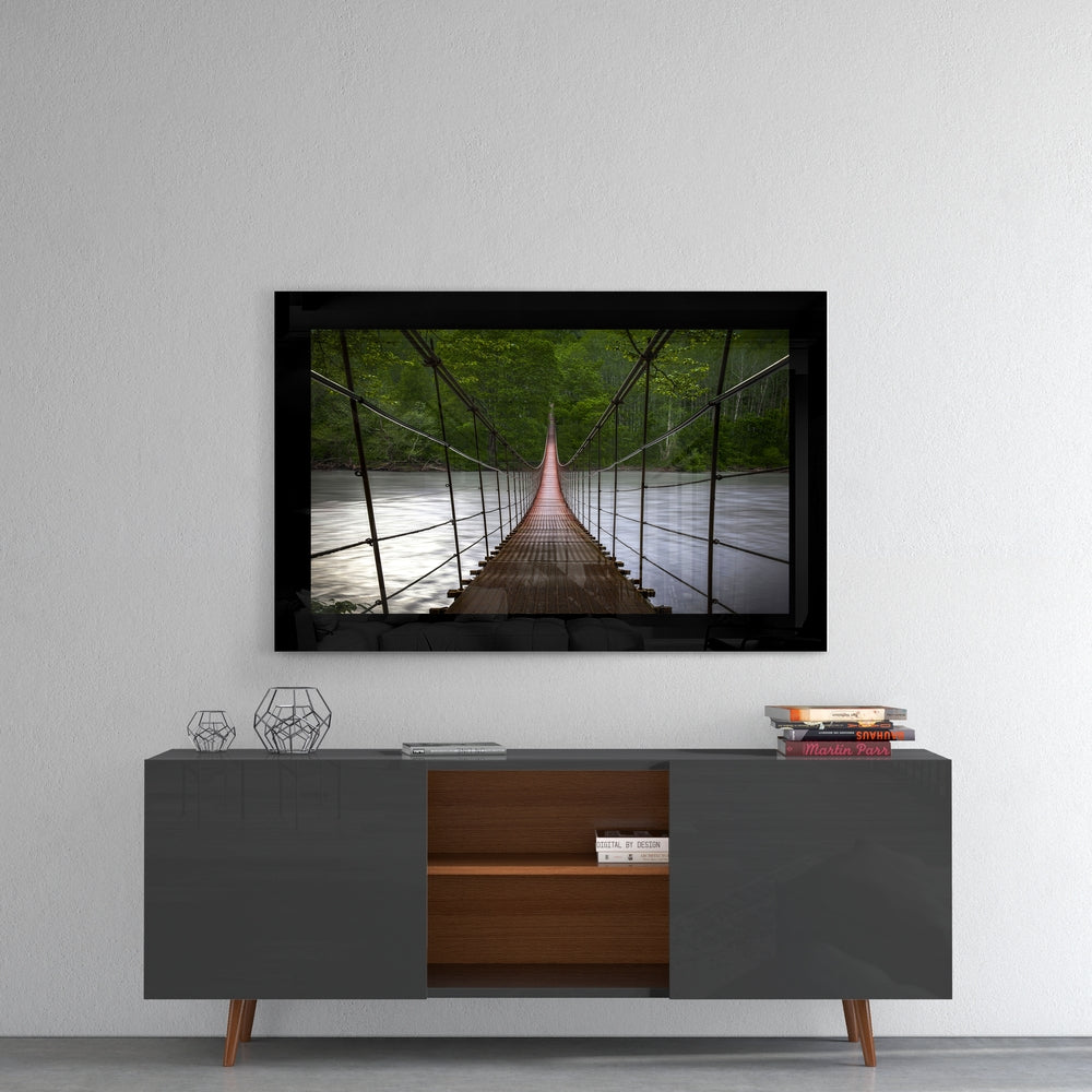 Framed glass wall art depicting a wooden suspension bridge stretching toward a river surrounded by green forest canopy, displayed above a mid-century modern credenza.