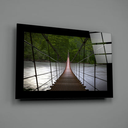 Modern glass wall art mounted on grey wall displaying a symmetrical suspension bridge spanning a river through green forest.
