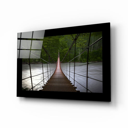 Black-framed glass wall art displaying a perspective view down a wooden suspension bridge over a river surrounded by green forest.
