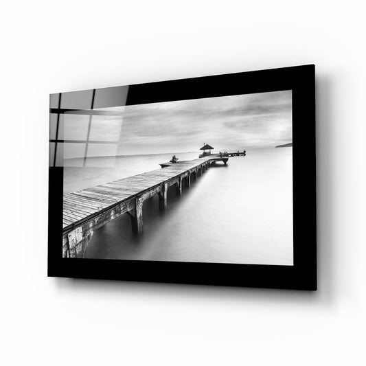 Black-framed glass wall art displaying a black and white photograph of a weathered pier extending into calm water with a pavilion structure visible at the end.