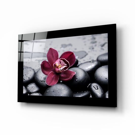 Black-framed glass wall art displaying a burgundy orchid flower on wet gray spa stones, mounted at an angle showing the frame's depth and shadow.