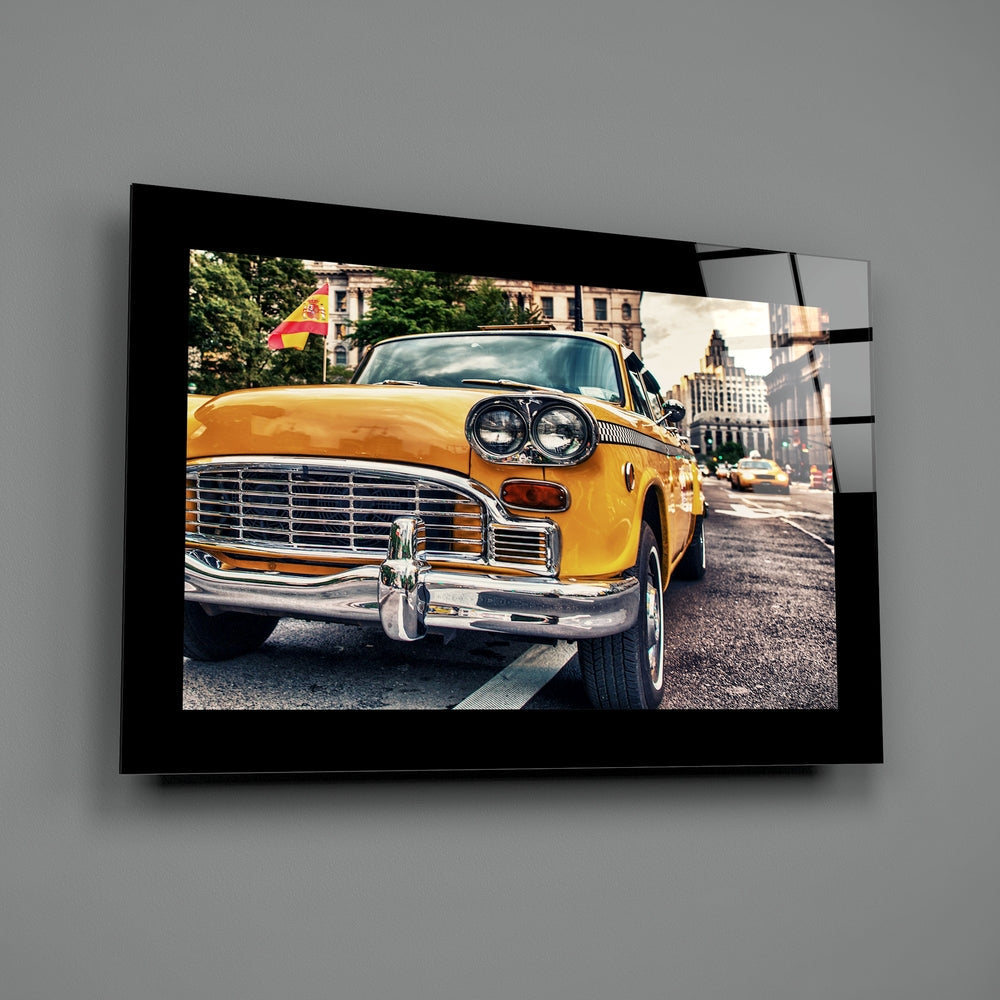 Black-framed glass wall art displaying a bright yellow vintage taxi cab on a city street, mounted on gray wall.