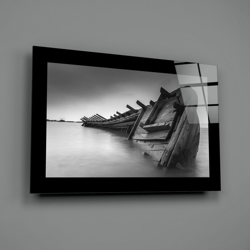 Black-framed glass wall art displaying a monochrome photograph of deteriorated wooden boat wreckage partially submerged in still water.