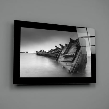 Black-framed glass wall art displaying a monochrome photograph of deteriorated wooden boat wreckage partially submerged in still water.