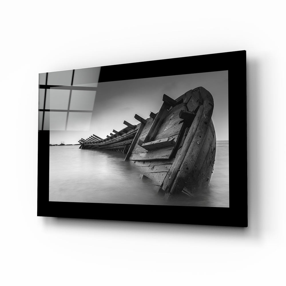 Black-framed glass wall art displaying a black and white photograph of deteriorated wooden boat wrecks partially submerged in calm water, shown at an angled perspective.
