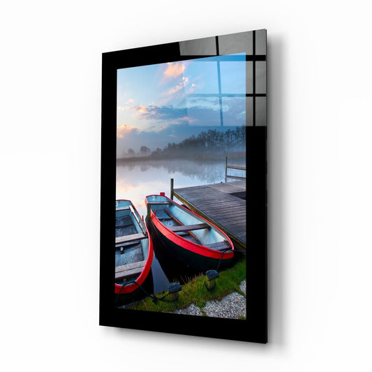 Black-framed glass wall art displaying red and white rowboats docked at a wooden pier beside a misty lake at dawn.