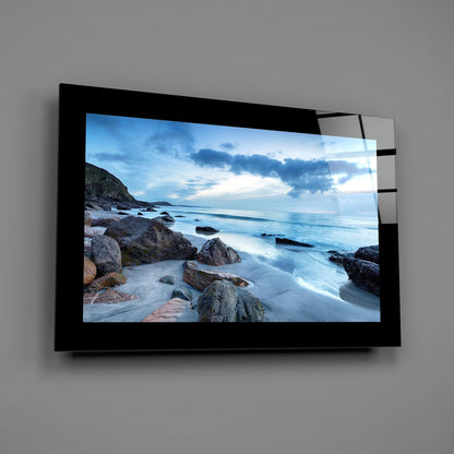 Black-framed glass wall art mounted on gray wall displaying rocky coastal beach scene with blue ocean and sky.