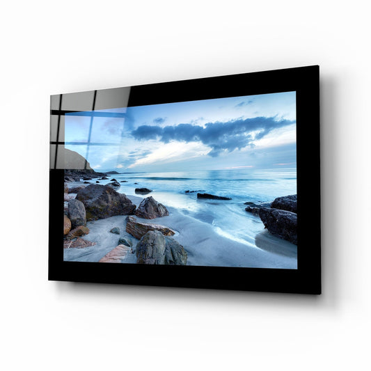 Black-framed glass wall art displaying a coastal seascape with rocks, beach, and sunset sky mounted at an angle.
