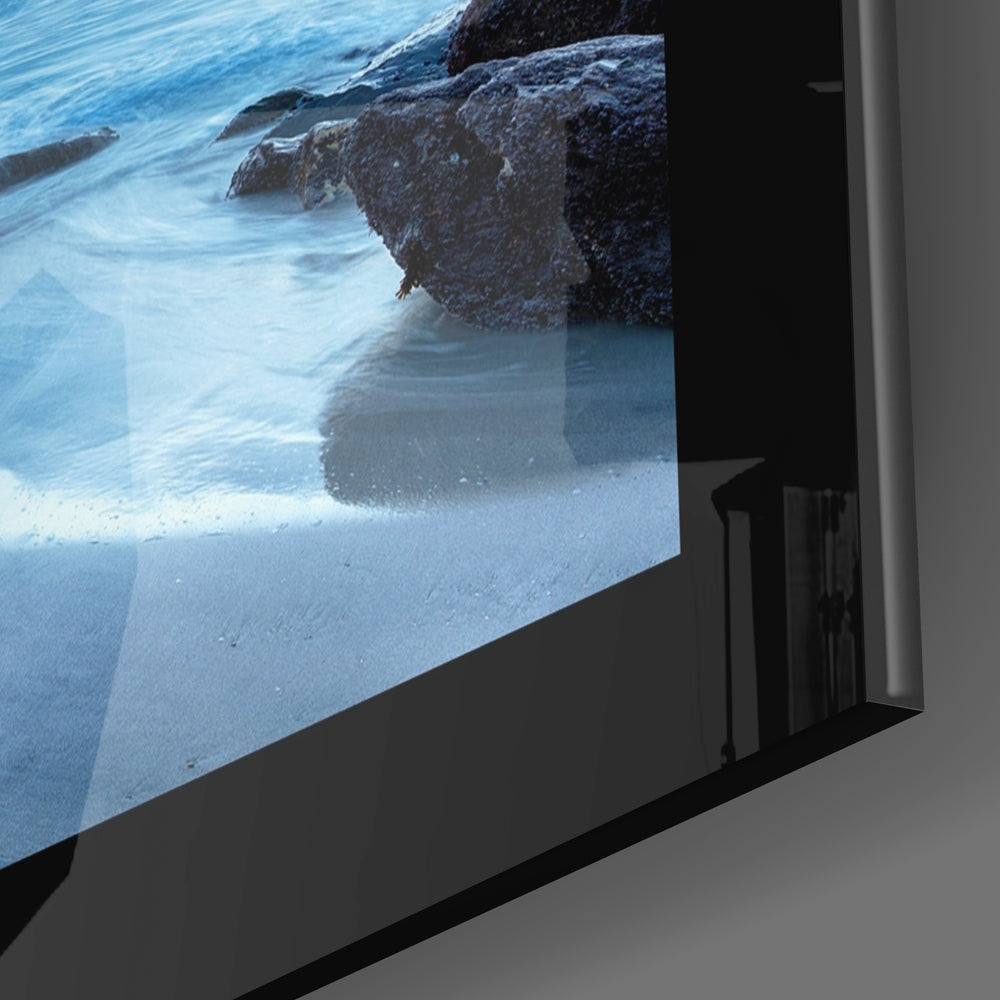 Glass wall art panel displaying coastal scene with rocky outcrop, turquoise water, and white sand beach, mounted at angle.