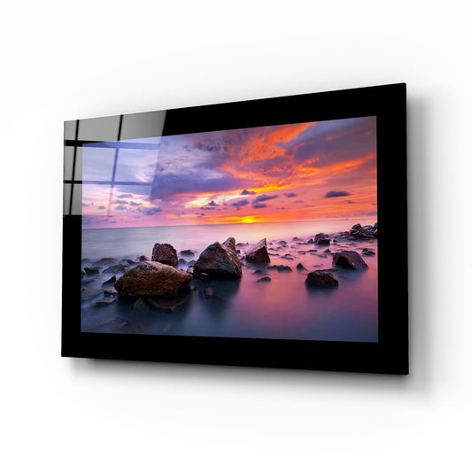 Black-framed glass wall art displaying a rocky coastline sunset with orange and purple sky, mounted at an angle showing reflective surface.