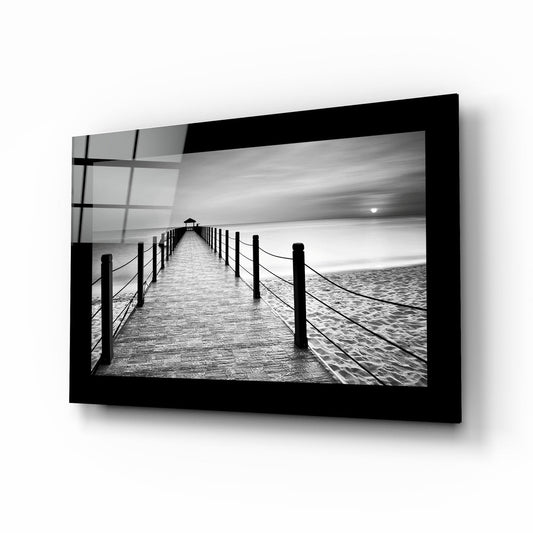 Black-framed glass wall art displaying a black and white photograph of a wooden pier with rope railings extending toward the horizon over calm water.