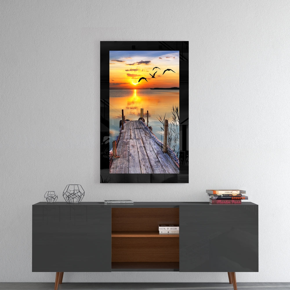 Framed glass wall art hanging above a mid-century console table, depicting a wooden dock extending toward a golden sunset with flying birds.