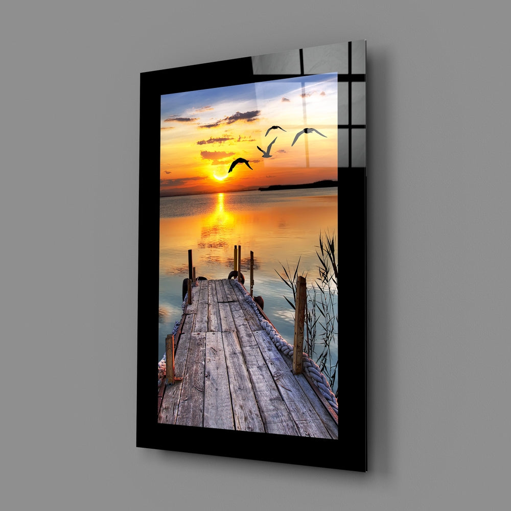 Black-framed glass wall art displaying a wooden dock extending toward a golden sunset with flying birds and water reflections.