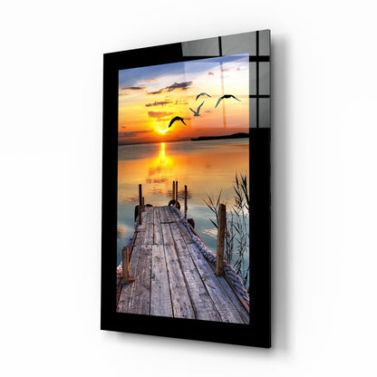 Black-framed glass wall art displaying a wooden dock perspective leading to a golden sunset with flying birds over water.