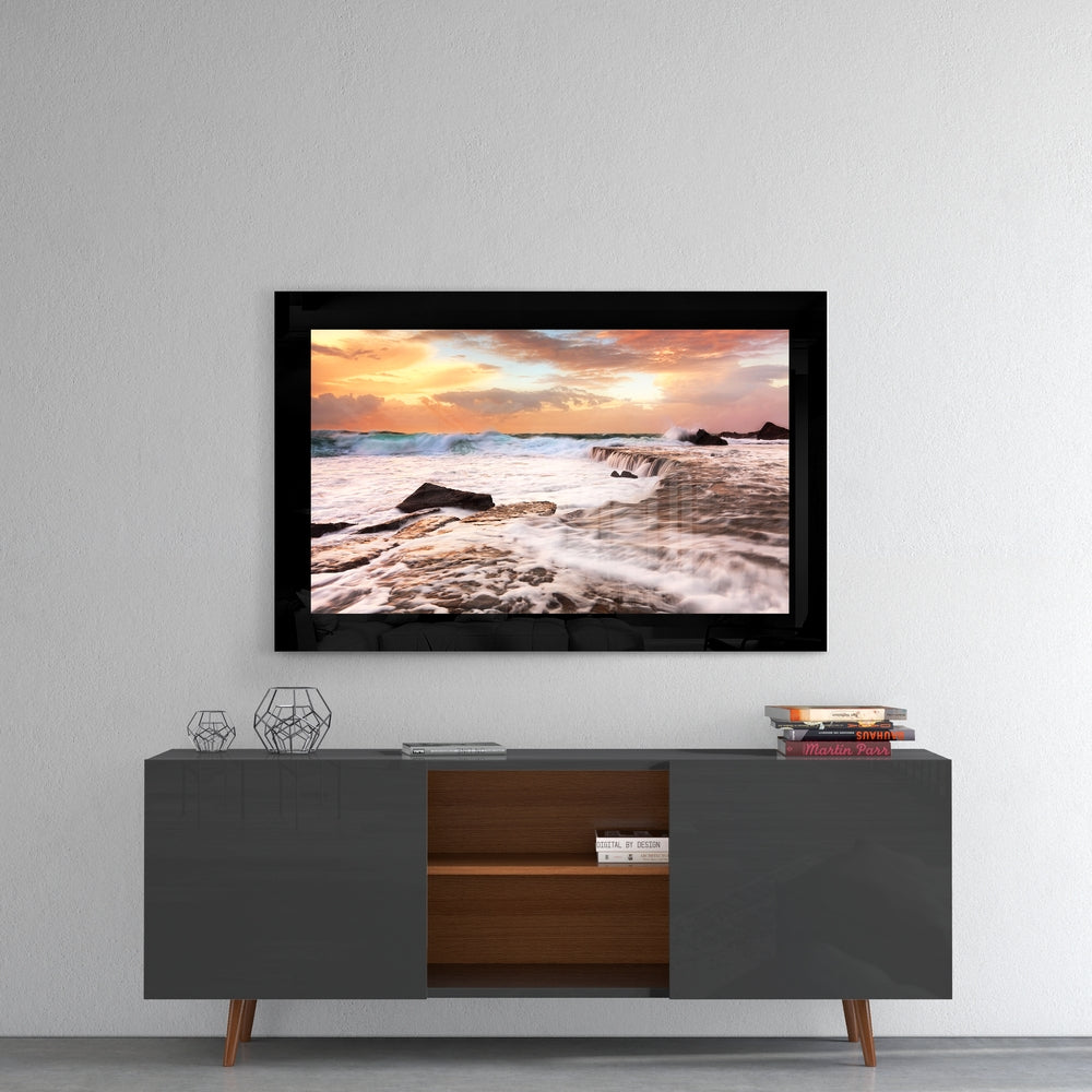 Framed glass wall art displaying a sunset beach scene with waves and rocks, mounted above a mid-century modern TV console.