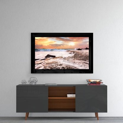 Framed glass wall art displaying a sunset beach scene with waves and rocks, mounted above a mid-century modern TV console.