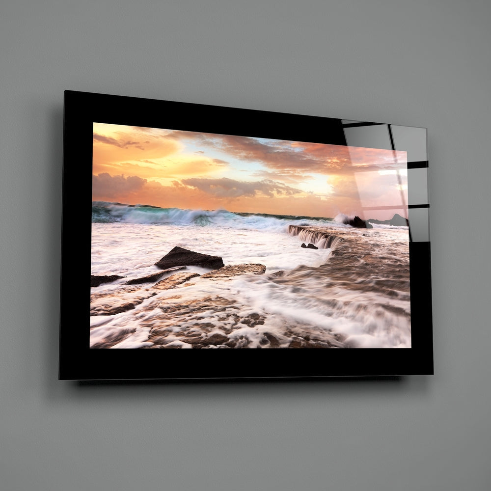 Black-framed glass wall art displaying a beach sunset with golden sky, waves, and rocks mounted on gray wall.