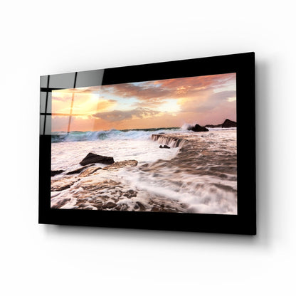 Black-framed glass wall art displaying a beach sunset with waves, rocks, and golden sky, mounted at an angle.