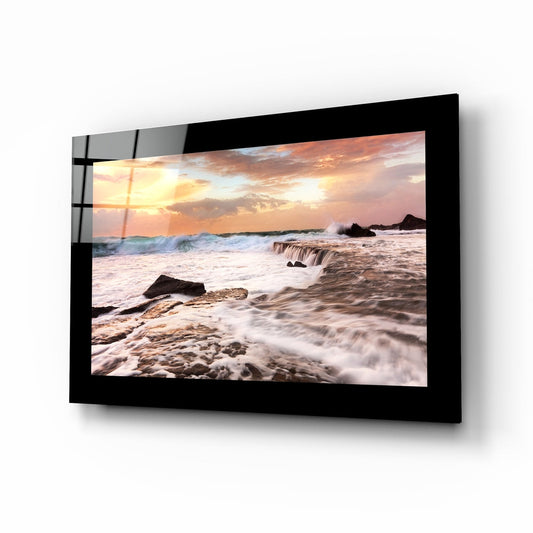 Black-framed glass wall art displaying a beach sunset with waves, rocks, and golden sky, mounted at an angle.