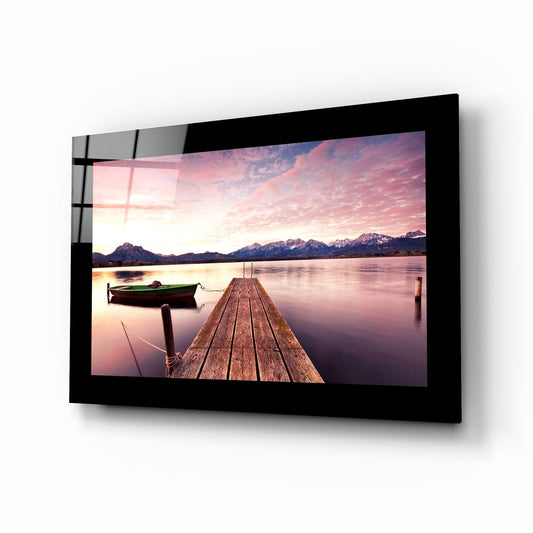 Black-framed glass wall art displaying a wooden dock extending into a pink-sunset lake with snow-capped mountains and a green boat, shown at an angled perspective.