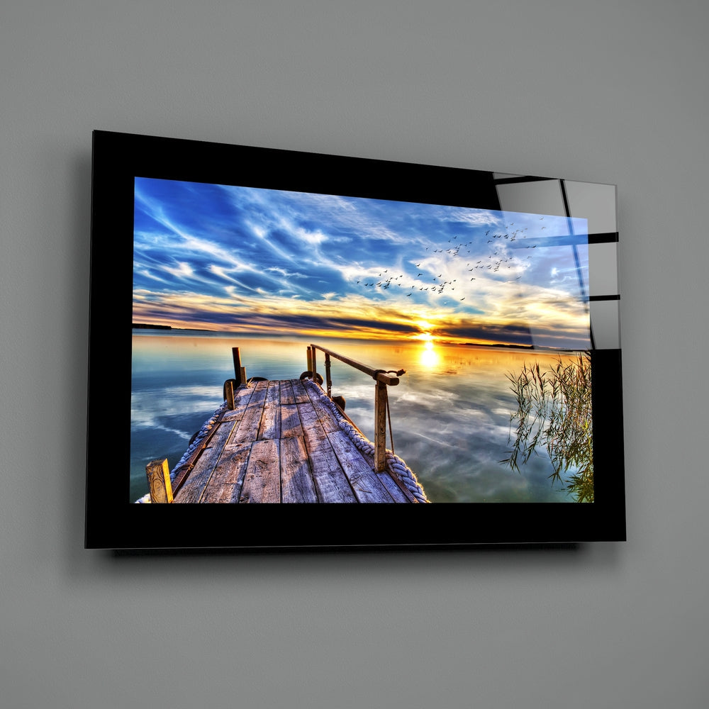 Black-framed glass wall art mounted on gray wall displaying wooden pier extending toward golden sunset over calm water with birds flying in blue sky.