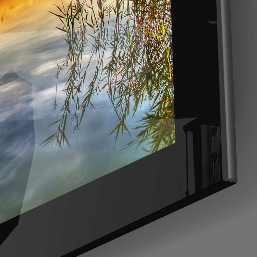 Angled view of framed glass wall art displaying weeping willow branches overhanging calm water at golden sunset.