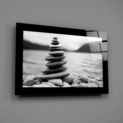 Black-framed glass wall art displaying stacked stones on a beach in monochrome, mounted on gray wall.