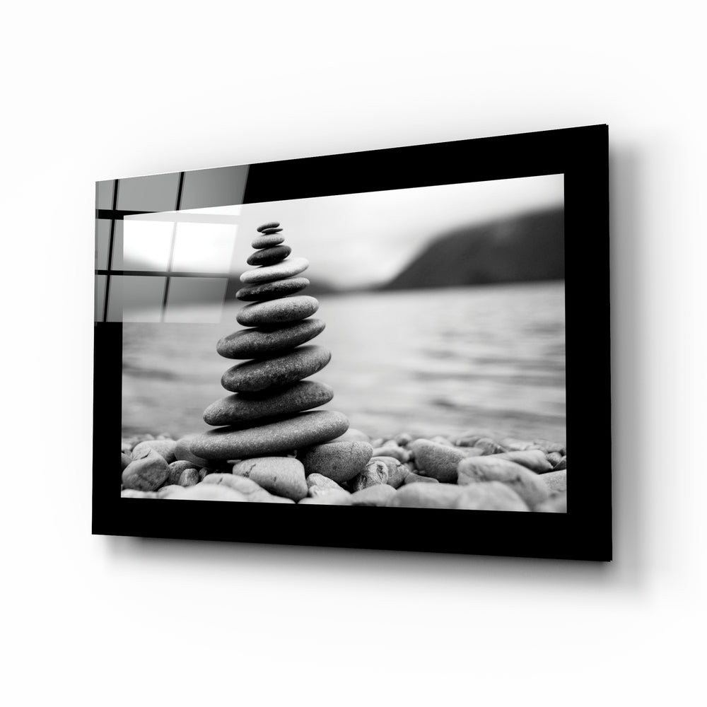 Black-framed glass wall art displaying a black and white photograph of stacked stones on a beach with blurred water background.