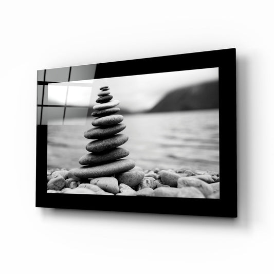 Black-framed glass wall art displaying a black and white photograph of stacked stones on a beach with blurred water background.