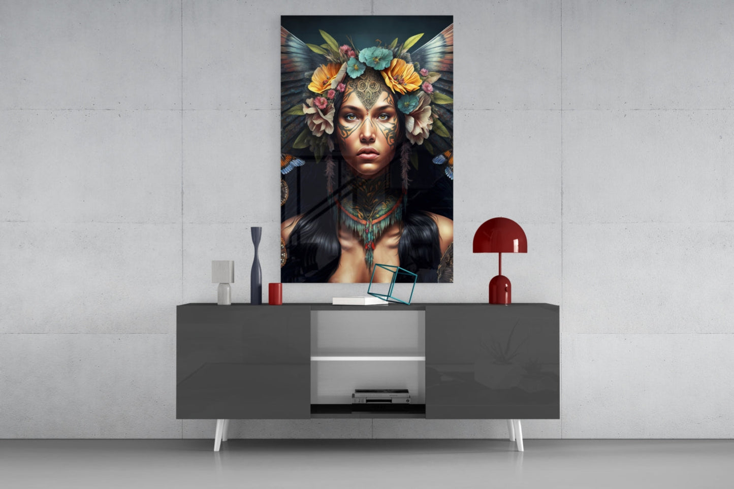 Glass wall art depicting a woman with ornate facial tattoos, colorful flowers, and dark wings, displayed above a modern gray console table with decorative objects.