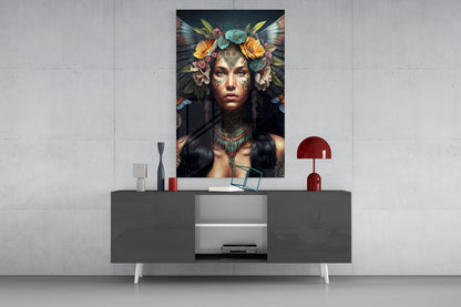 Glass wall art depicting a woman with ornate facial tattoos, colorful flowers, and dark wings, displayed above a modern gray console table with decorative objects.