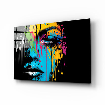 Angled perspective view of a glass wall art panel featuring a colorful pop art portrait with vibrant blue, yellow, and magenta paint drips on black background.