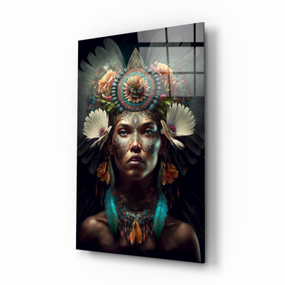 Glass wall art panel displayed at an angle showing a portrait of a woman with intricate facial tattoos, elaborate floral headdress, and feathered adornments against a dark background.