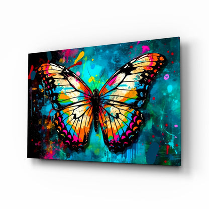 Colorful butterfly glass wall art with vibrant orange, yellow, magenta, and teal wings against a dark turquoise splattered background, displayed at a three-quarter angle.