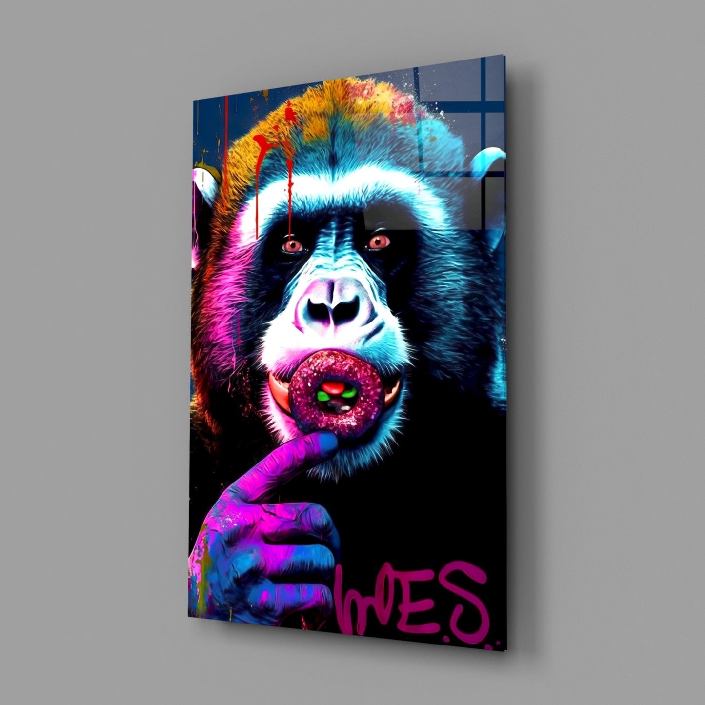 Angled view of vibrant pop art glass wall panel featuring colorful chimpanzee with glowing eyes holding lollipop, magenta text at bottom, mounted on gray wall.