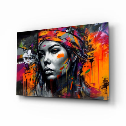 Angled view of a glass wall art panel featuring a monochrome woman's face with vibrant multicolored graffiti-style paint splatters in orange, pink, and purple.