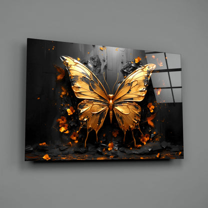 Golden butterfly glass wall art displayed at an angle against gray wall, showing luminous amber insect with orange flower petals and dark architectural background.