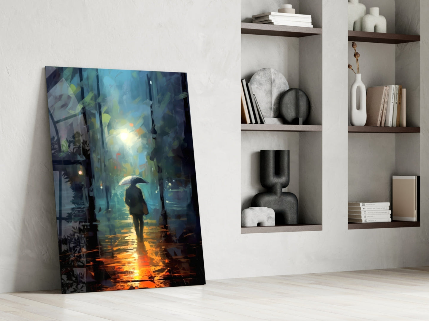 Glass wall art leaning against white wall showing figure with umbrella in rain-lit street scene in blue and orange tones, displayed beside white shelving unit with decorative objects and books.