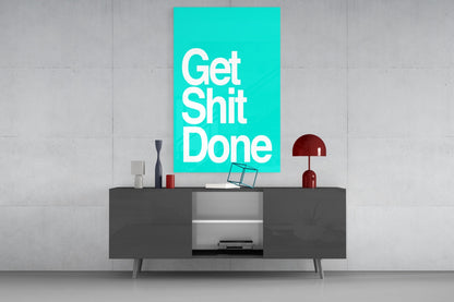 Turquoise glass wall art with "Get Shit Done" text displayed above dark sideboard with decorative objects and lamp.