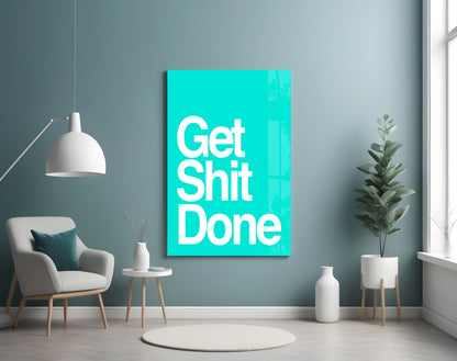 Bright turquoise glass wall art displaying "Get Shit Done" in white text, mounted in a modern living room setting with gray walls, furniture, and potted plants.