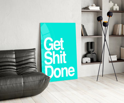 Turquoise glass wall art with white "Get Shit Done" text displayed in modern living room next to black leather sofa and shelving unit.