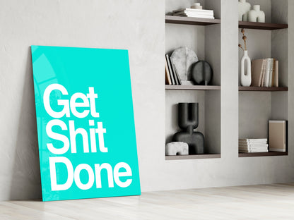 Turquoise glass wall art panel leaning against white wall displaying "Get Shit Done" in white text, with shelving and decor visible behind.