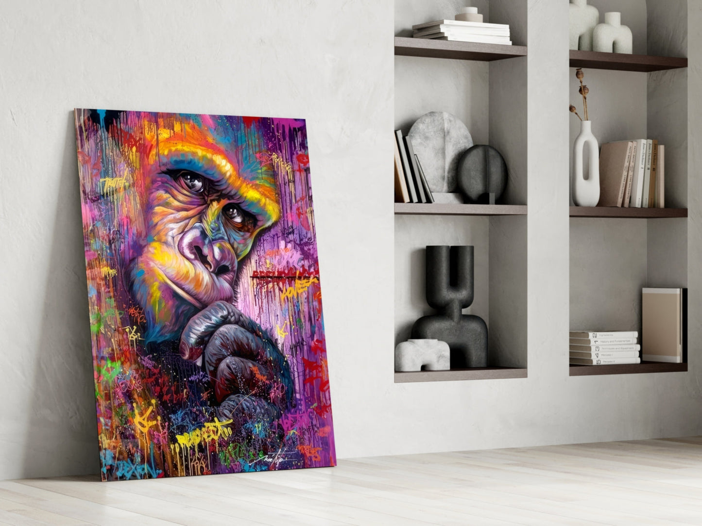 Colorful street art primate portrait leaning against white wall beside minimalist shelving with books and decorative objects.