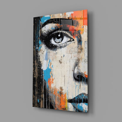 Angled view of glass wall art featuring a stylized eye portrait with orange, blue, and cream colors on distressed background.