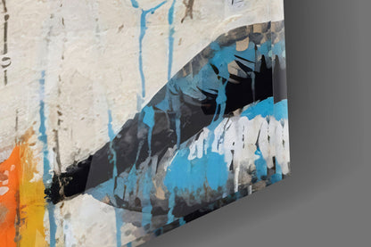 Close-up angled view of glass wall art showing blue, white, and black painted portrait with orange and yellow accent, mounted on white textured wall.