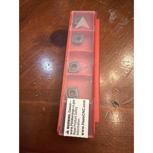 Red plastic storage case with five compartments containing gray carbide inserts, warning label visible on front, photographed from above on wooden surface.