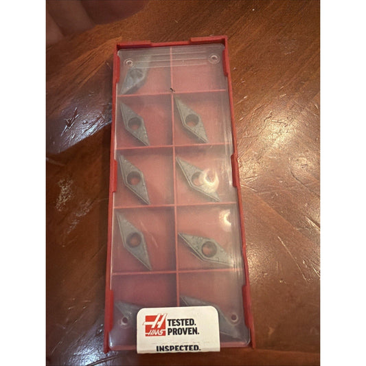 Red plastic storage case containing nine metallic carbide turning inserts arranged in a 2x5 grid with Haas "Tested. Proven. Inspected." label on white sticker.